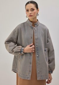 Svetlo siva jeans jakna z ovratnikom za stoječe nošenje, gumbnim zapenjanjem spredaj in nabranimi manšetami na rokavih. Ima sproščen kroj in stranske žepke.