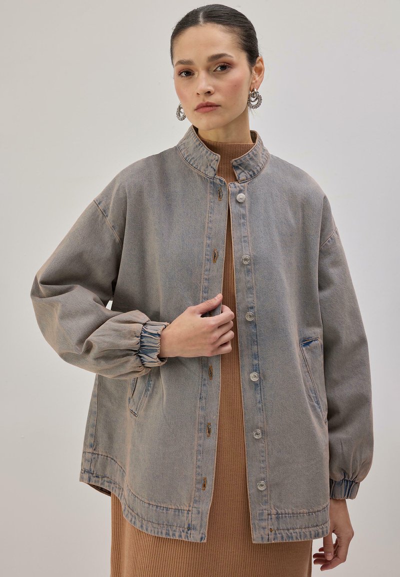 Svetlo siva jeans jakna z ovratnikom za stoječe nošenje, gumbnim zapenjanjem spredaj in nabranimi manšetami na rokavih. Ima sproščen kroj in stranske žepke.