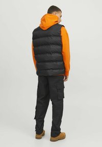 Sorte puffer vest over en orange hoodie med lynlås, iført cargo-bukser og brune støvler, set fra ryggen.