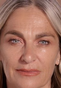 Nahaufnahme eines Gesichts mit heller Haut, grauem Haar, blauen Augen, leichten Falten und natürlichem Make-up, einschließlich eines subtilen Lippenfarbtons.
