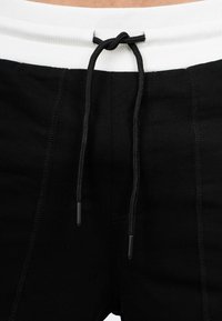 Pantalons de survêtement noirs avec une taille côtelée blanche, ornés d'un cordon de serrage noir. Les détails de couture visibles rehaussent le design.