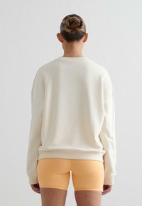 Felpa crema con spalle scese, polsini e orlo a coste; abbinata a pantaloni shorts arancioni aderenti. Design minimalista, superficie liscia.