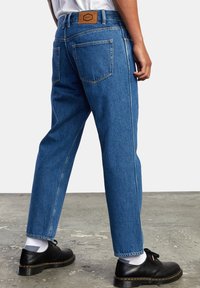 Pantalones de mezclilla azul con un corte relajado, cinco bolsillos y costuras en contraste. Parche de cuero marrón en la cinturilla, combinados con zapatos negros.