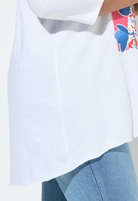 Witte katoenen T-shirt met een ronde zoom, versierd met een bloemenpatroon in blauw en roze op de voorkant. Draagt samen met lichtblauwe jeans.