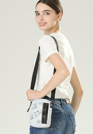 Giovane donna con capelli scuri raccolti, che indossa una maglietta bianca e jeans blu, porta una borsa a tracolla bianca con testo nero e cerniere.