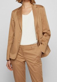 Kobieta ubrana w kamelowy, dopasowany blezer i pasujące spodnie, z białą, prążkowaną, bezrękawnikową bluzką, z ręką w kieszeni, na jasnoszarym tle.