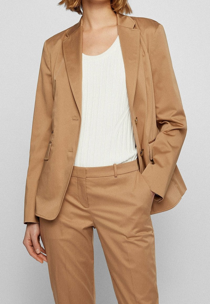 Kobieta ubrana w kamelowy, dopasowany blezer i pasujące spodnie, z białą, prążkowaną, bezrękawnikową bluzką, z ręką w kieszeni, na jasnoszarym tle.