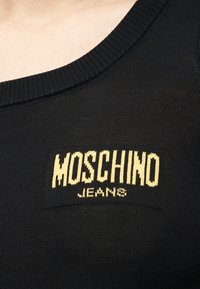 Κοντινό πλάνο στο λαιμό μαύρου πλεκτού με κίτρινο μπαλώμα λογότυπου «MOSCHINO JEANS» στο στήθος.