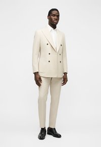 Mand iført en beige dobbeltradet jakkesæt med en hvid skjorte og sorte pæne sko, stående mod en ensfarvet hvid baggrund.