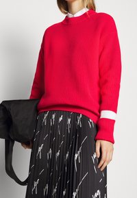 Pull à côtes rouge avec un col rond et des accents de rayures blanches sur les poignets, associé à une jupe plissée noire avec des motifs de texte blancs.