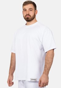T-shirt bianco a maniche corte con scollatura rotonda, caratterizzato da un'etichetta di marca sull'orlo e una texture liscia simile al cotone.