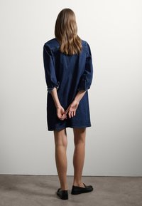 Denim shirtjurk in donkerblauw, met lange mouwen die bij de manchetten opgerold zijn, een relaxte pasvorm en een rechte zoom. Gecombineerd met zwarte platte schoenen.