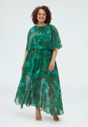 Frau lächelnd, trägt ein grünes, blattgemustertes, durchsichtiges Maxikleid mit geschichtetem Oberteil und goldenen Riemchenabsätzen, steht vor einem schlichten weißen Hintergrund.
