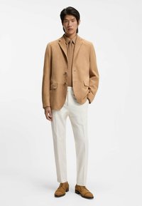 Mann trägt einen hellbraunen Wildlederblazer, dazu passende Krawatte und Schuhe, beiges Hemd und weiße Hose, steht vor weißem Hintergrund.