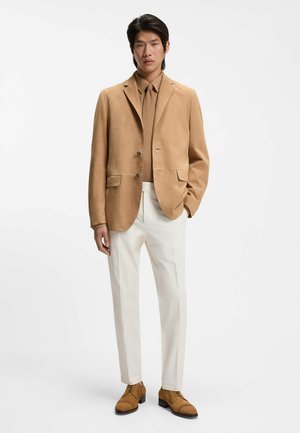 Man draagt een zandkleurig suède blazer, bijpassende stropdas en schoenen, beige overhemd en witte broek, staande tegen een witte achtergrond.