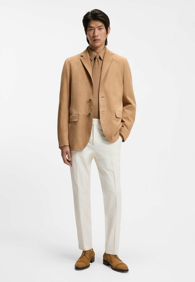 Mann trägt einen hellbraunen Wildlederblazer, dazu passende Krawatte und Schuhe, beiges Hemd und weiße Hose, steht vor weißem Hintergrund.