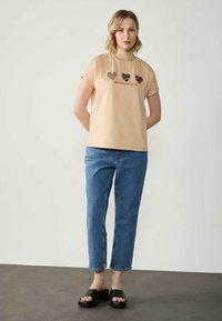 Beige cotton t-shirt with short sleeves, featuring three heart graphics in various patterns, paired with blue cropped denim jeans and black sandals.