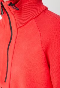 Gros plan d'une veste polaire rouge avec un col montant côtelé, une fermeture éclair noire et un panneau d'épaule en tricot texturé.
