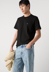 Black crew-neck t-shirt in cotton, featuring a small black label on the chest. Worn with light blue denim jeans, one hand in pocket.