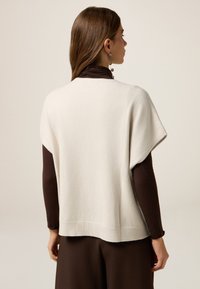 Lichtbeige oversized trui met korte mouwen, geribbeld einde en een zachte textuur. Draagt over een bruine coltrui, wat een gelaagde stijl laat zien.
