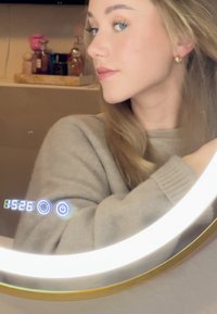 Jeune femme avec de longs cheveux blonds et un pull gris ajustant ses cheveux dans un miroir rond éclairé affichant l'heure numérique et des commandes tactiles.
