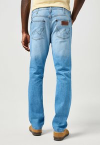 Jeans de mezclilla azul claro con un ajuste recto, que cuentan con una etiqueta de cuero marrón, bolsillos traseros con costuras en zigzag y una textura desgastada.