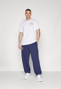 Man standing with right hand in pocket, wearing white Under Armour t-shirt, navy sweatpants, and white sneakers against plain light background.