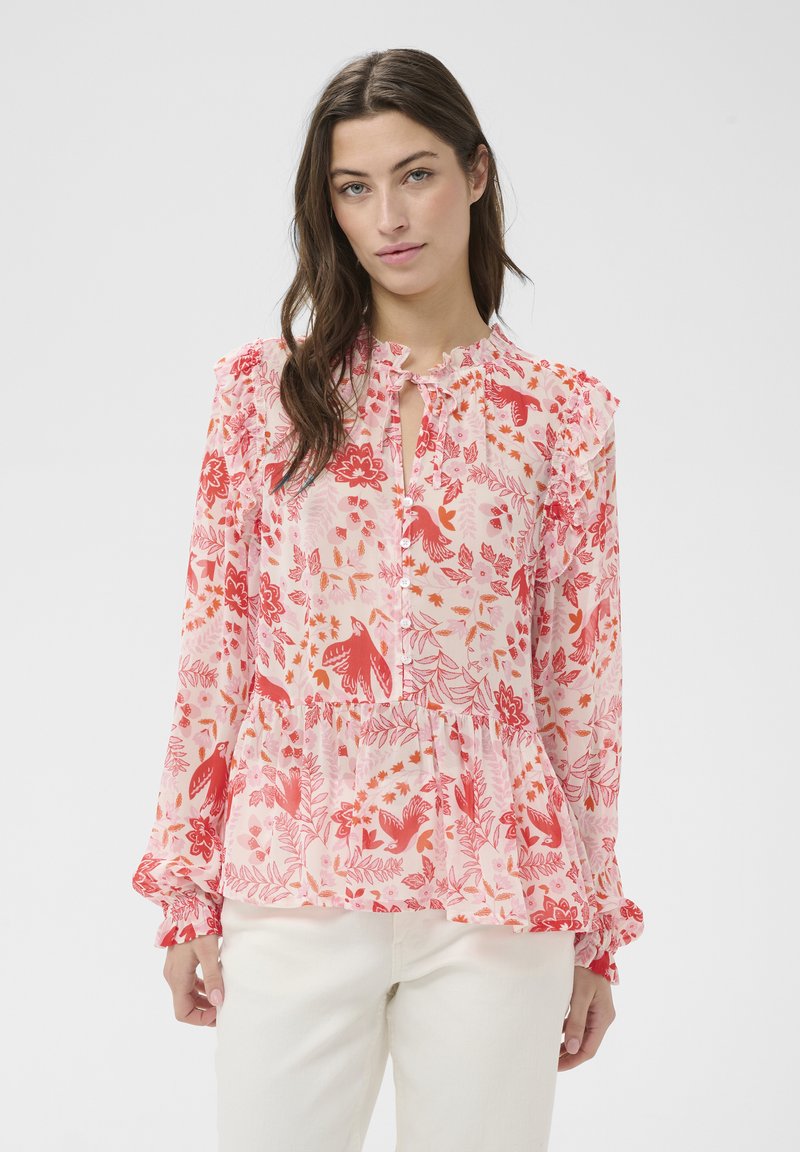 Kobieta ubrana w różową bluzkę z czerwonymi kwiatowymi i ptasimi wzorami, z falbanami na ramionach i mankietach, w połączeniu z białymi spodniami, stojąca na jednolitym tle.