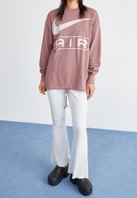 Př oversized růžová mikina s bílým logem Nike, spárovaná s bílými pruhovanými rozšířenými kalhotami a černými robustními dřeváky s ventilačními otvory.