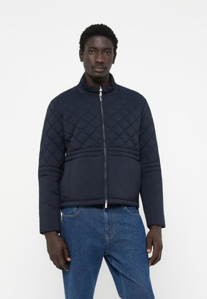 Navy quilted jacket with a high collar, zip closure, and horizontal seam details. Ribbed trim at the hem. Paired with blue jeans.
