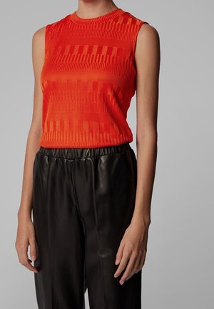 Sleeveless red textured top with geometric patterns paired with high-waisted black leather pants on a person against a gray background.