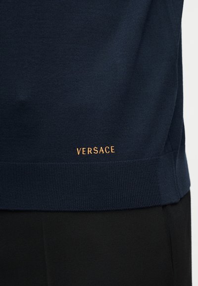 Ourlet de pull en maille bleu foncé avec logo "VERSACE" brodé en or au-dessus d'un pantalon noir.