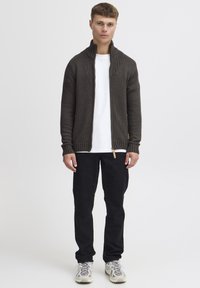 Cardigan a maglia marrone scuro con collo alto a coste, polsini a coste e una cerniera con accentuazione in legno naturale. Indossato sopra una t-shirt bianca e pantaloni neri.