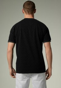 Camisa corta negra con un patrón geométrico texturizado, cuello redondo y puños acanalados en contraste, vista desde la parte de atrás. Pantalones blancos visibles.
