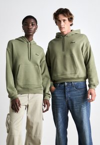 Dos personas vestidas con sudaderas verde oliva con un bolsillo frontal y un logo bordado. Un par de pantalones beige y un par de jeans azules.