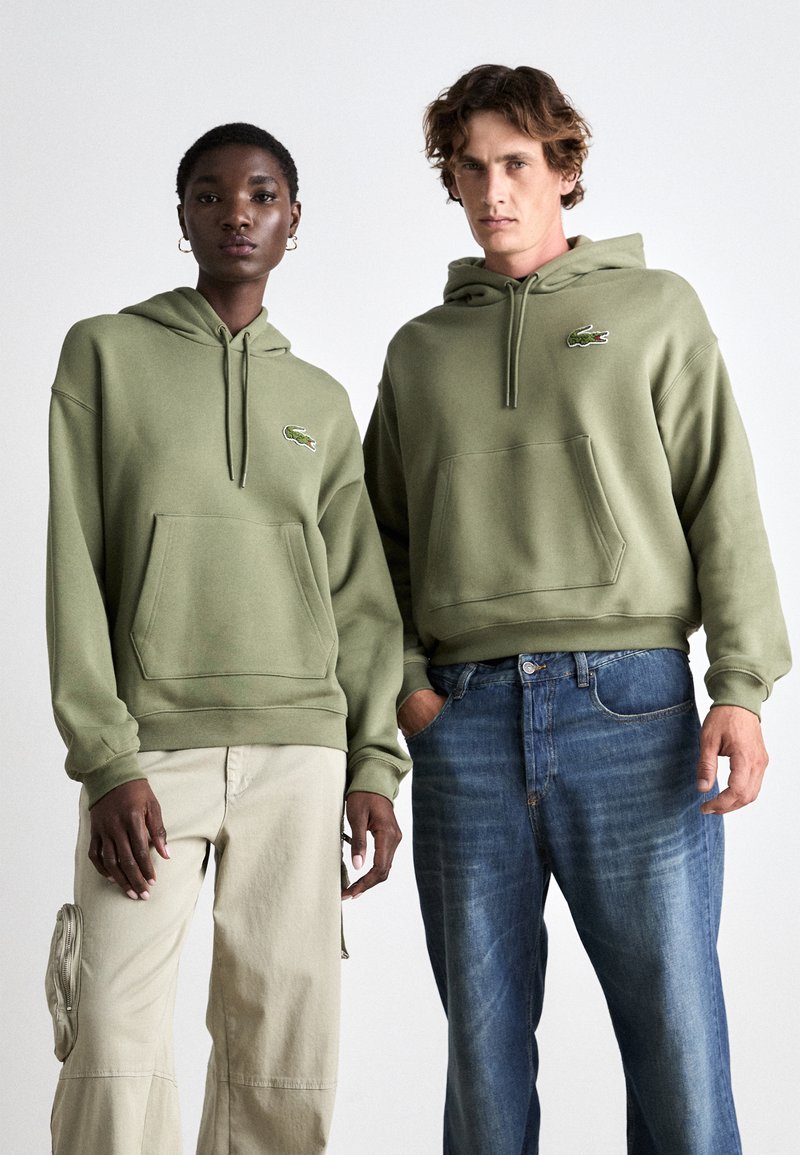 Dos personas vestidas con sudaderas verde oliva con un bolsillo frontal y un logo bordado. Un par de pantalones beige y un par de jeans azules.