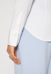 Main d'une femme portant des bagues en argent reposant à côté, vêtue d'une chemise blanche à manches longues avec poignets boutonnés et d'un pantalon bleu clair.