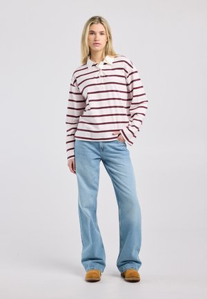 Jonge vrouw die staat met haar hand in haar zak, gekleed in een witte en bordeaux gestreepte polo met lange mouwen, lichtblauwe spijkerbroek en bruine schoenen, neutrale achtergrond.