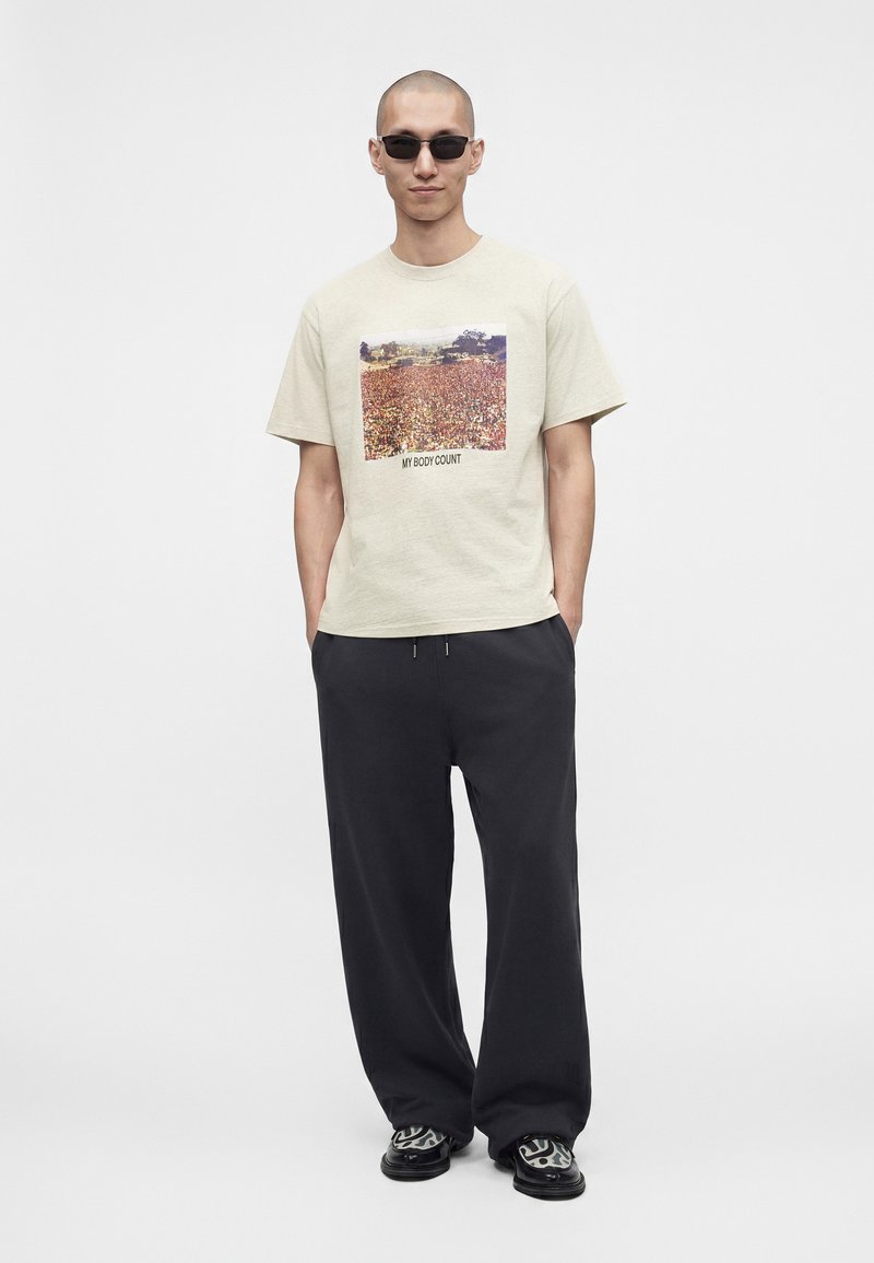 Man wearing sunglasses, a beige t-shirt with a crowd photo and text "MY BODY COUNT," loose black pants, and patterned black shoes.