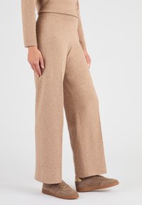 Pantaloni beige a gamba larga realizzati in tessuto morbido, con vita alta e una texture liscia. Abbinati a sneakers in suede marroni.