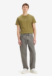 Camiseta de algodón verde oliva con un pequeño logotipo blanco, combinada con jeans grises de corte holgado y zapatillas blancas. Diseño minimalista.
