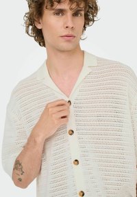White short-sleeve shirt with a textured, open knit pattern; features a collared neckline and large brown buttons along the front.