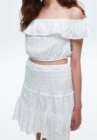 Femme portant un haut court blanc à épaules dénudées en tissu œillet et une jupe assortie, avec un tatouage visible sur le bras et des cheveux bruns lâchés.