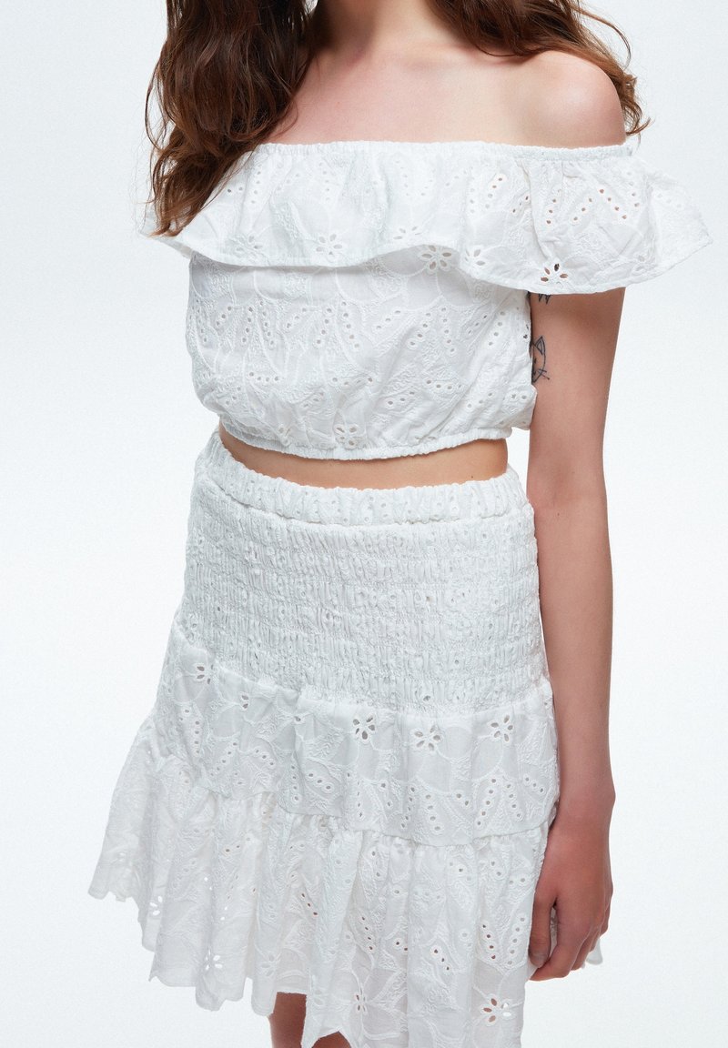 Femme portant un haut court blanc à épaules dénudées en tissu œillet et une jupe assortie, avec un tatouage visible sur le bras et des cheveux bruns lâchés.