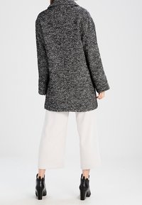 Cappotto grigio e bianco a motivo houndstooth con colletto, maniche lunghe e taglio dritto, abbinato a pantaloni larghi color crema e stivali alla caviglia.