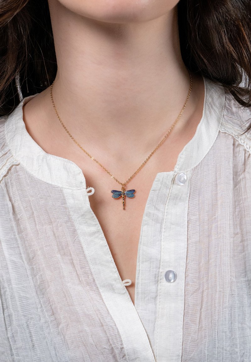 Guldhalsband med ett fint hänge föreställande en blå och svart trollslända, buren av en person i en vit texturerad blus.