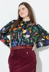 Una donna con long capelli castani indossa una blusa nera a fiori con fiori colorati e chiavi, abbinata a una gonna bordeaux e una cintura intrecciata.