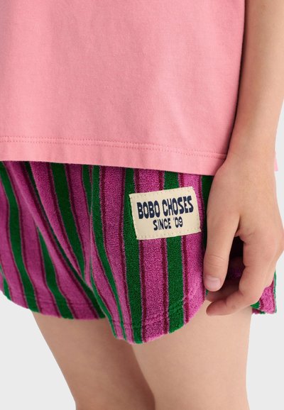 Enfant portant un haut rose et un short rayé rose et vert avec une étiquette "BOBO CHOSES SINCE '09" près de la main.