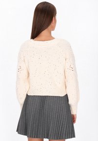 Femeie cu părul lung castaniu, purtând un pulover crem din tricot cu găuri pe mâneci și o fustă plisată gri închis, văzută din spate.