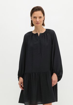 Vrouw draagt een losse zwarte jurk met lange mouwen, een geplooide halslijn en een gelaagde rok, staand tegen een effen lichte achtergrond.